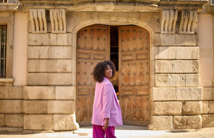 A woman in a pink suit smiles while walking past an old wooden door of a stone building.
