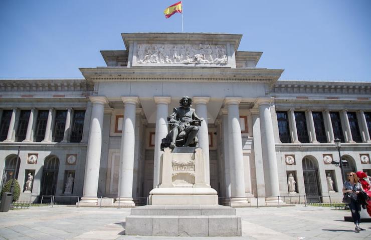 A view of the Prado Museum entrance featuring a statue in the foreground and the Spanish flag above.