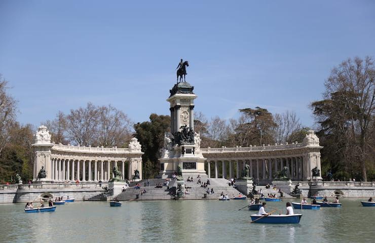 A lively scene of people boating and enjoying a sunny day around a large monument with columns and a statue in a public park.