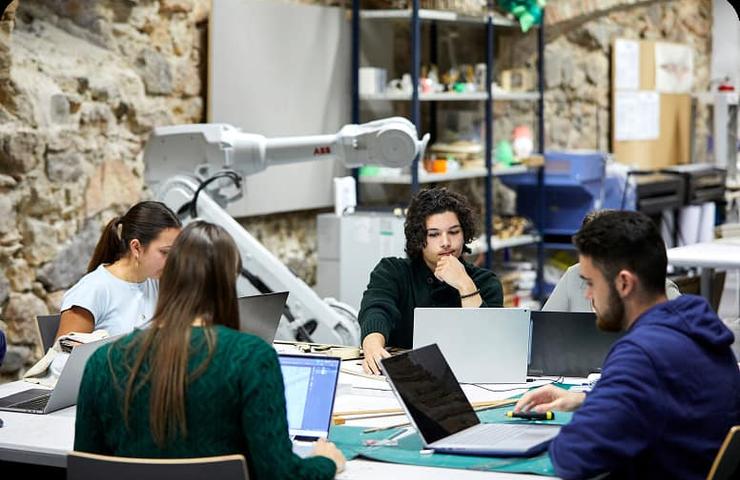 A group of students collaborates on a project at a workspace with computers and a robotic arm in the background.