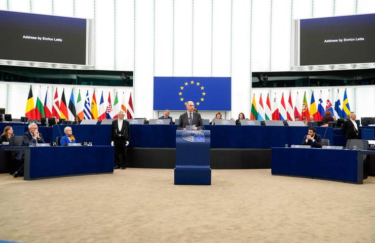 A formal address being delivered at the European Parliament with a backdrop of various national flags.