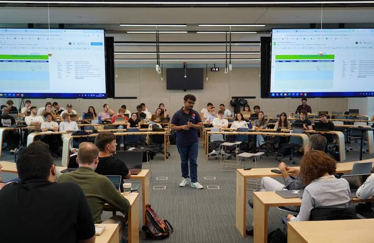 A speaker engages a large audience in a modern classroom setting with screens displaying content.