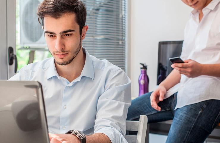 Two men in a modern office setting, one focused on a laptop and the other using a smartphone.