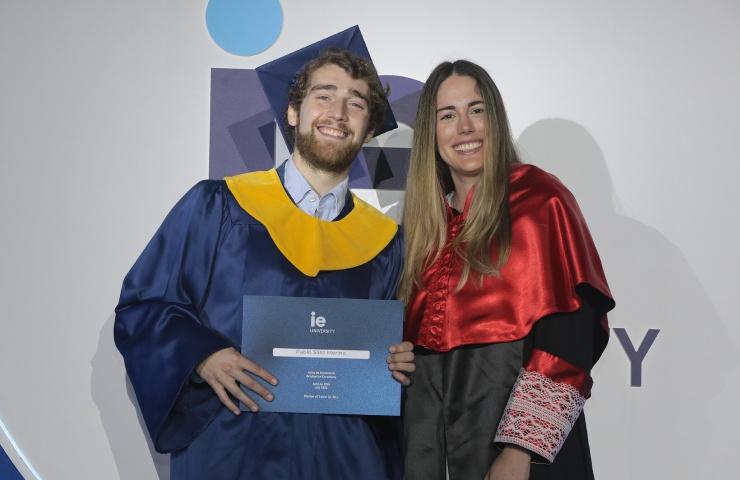 A young man and woman in graduation robes smiling and holding a diploma at a ceremony.