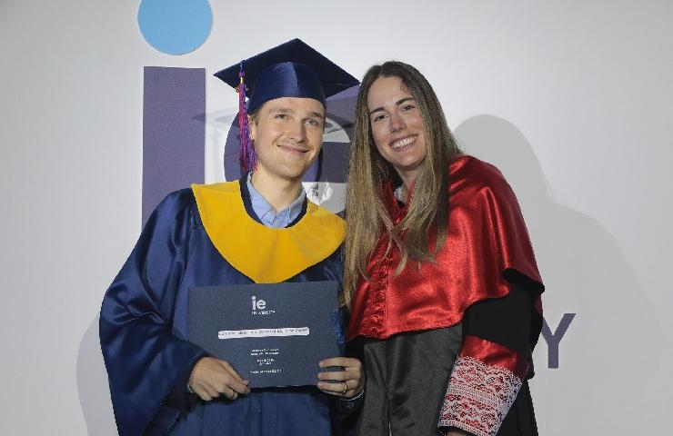 A young man in a blue and yellow graduation cap and gown holding a diploma, standing next to a young woman in a red graduation stole.