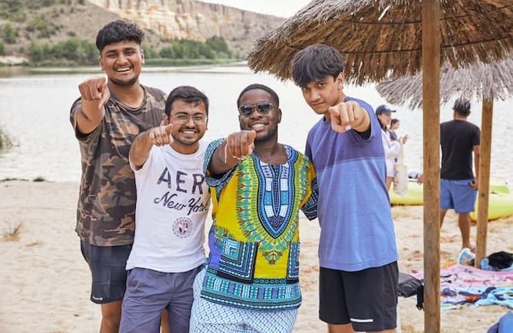 Four young men smiling and pointing towards the camera at a lakeside beach.