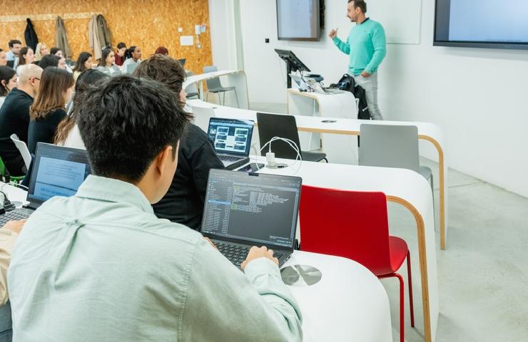 A classroom scene with students seated at desks, focused on their laptops while a teacher presents at the front.