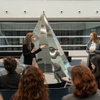 Two women are engaged in a discussion in front of a shiny pyramid sculpture in a modern space.