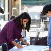 A woman is focused on drawing on a large sheet of paper at an outdoor table while a man watches.