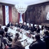 A formal meeting in an elegant room with a long table and attendees engaged in discussion.