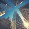 Looking up at towering skyscrapers against a clear blue sky with sunlight shining through.