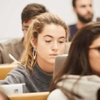A focused young woman using a laptop in a classroom full of students.