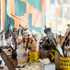 A cluttered artist's table filled with paint brushes, open paint cans, and various painting supplies in front of a colorful abstract painting.