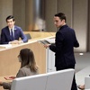 A man is speaking at a podium in a modern conference room with several attendees and a panel of two men and a woman.