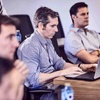 A group of men focused on laptops and listening during a business meeting in a modern office.