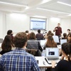 A lecturer explaining to students in a classroom equipped with laptops and a projector screen.