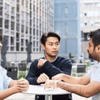 Three people having a conversation at a table outdoors, with modern buildings in the background.