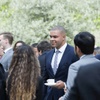 A man in a suit with a tie is smiling while holding a coffee cup, interacting with other people at an outdoor gathering.