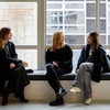Three women sitting together in a modern space, discussing something while looking out a window.