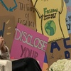 Two women seated in front of colorful protest signs, engaging in a discussion at a public event.