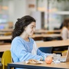 A young woman smiling as she uses a laptop in a cafeteria, with food and a drink on the table.