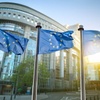 Three European Union flags flutter in front of a modern glass building on a sunny day.