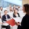 A woman is presenting to an audience in a seminar hall, holding a red folder and addressing attendees who are seated and listening attentively.