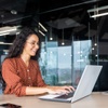 A woman in a polka dot shirt smiling and working on a laptop in a modern office environment.