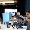 A young man assembles a robotic device at a workspace with various technology items in the background.