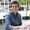 A smiling man working on a laptop at an outdoor table in a modern urban setting.