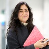 A smiling young woman holding a notebook and a phone in a modern office environment.