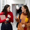 Two young women happily conversing while walking through a school corridor, one holding a notebook and the other a cup.