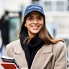 A smiling woman wearing a university cap and a beige coat holds books, standing outdoors.