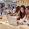 Two women collaborate over a laptop in a spacious co-working environment filled with people.
