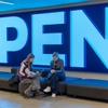 Two women are sitting on a bench in front of a large illuminated 'OPEN' sign.