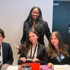 A group of four women in professional attire are seated at a table in a conference setting.