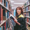 A graduate in a black robe holding a book in a library aisle, smiling at the camera.