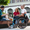 Three friends, one in a wheelchair, chatting and smiling in an outdoor urban setting.