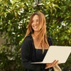 A young woman smiles while holding a laptop, standing outdoors surrounded by green foliage.