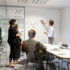 A team meeting in a modern office space with a person presenting on a whiteboard.