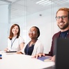 A group of three colleagues smiling and sitting at a table in a modern office.