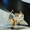 A man and a woman sitting and talking under a large, spiral staircase in a modern building.
