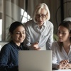 An older woman instructs two younger women using a laptop in an office setting.