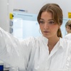 A woman in a laboratory is carefully adding a liquid from a pipette into a container.