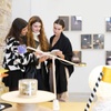 Three women examining a piece of fabric in an art gallery with various artworks displayed in the background.