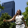 A young woman smiles while walking among trees with a modern building in the background.