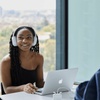 A young woman with headphones smiles while working on a laptop in a modern workspace.