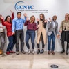 A group of nine diverse people standing in a line, smiling and posing with thumbs up in front of a wall that displays the logo and name of Glasgow Caledonian New York College.