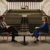 A formal interview setting with two people seated at a table on a stage in an empty auditorium.