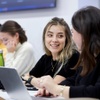 A group of students engaged in conversation and work at a table in a classroom setting.
